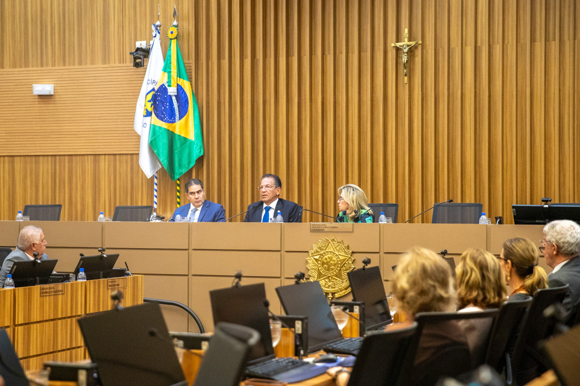 "Ministro Campbell fala ao microfone ao lado da Dra. Ana Paula e juiz Lizandro, sendo observado por membros da Corte no plenário"