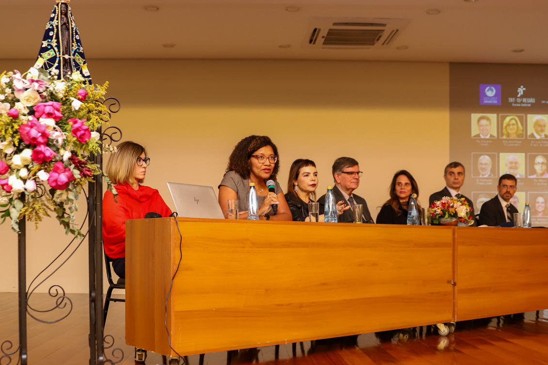 "Dra. Rosilene fala ao microfone na mesa alta ao lado de outros componentes do painel"