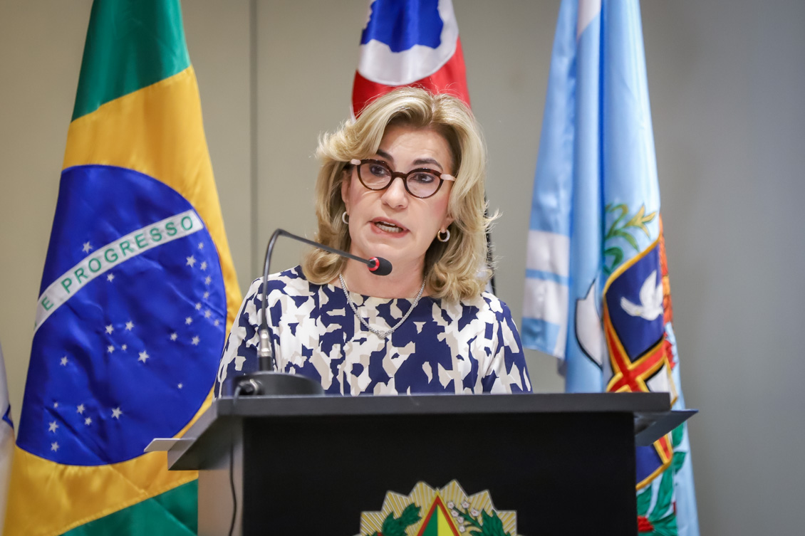 "Dra. Ana Paula está falando no púlpito em frente a duas bandeiras"