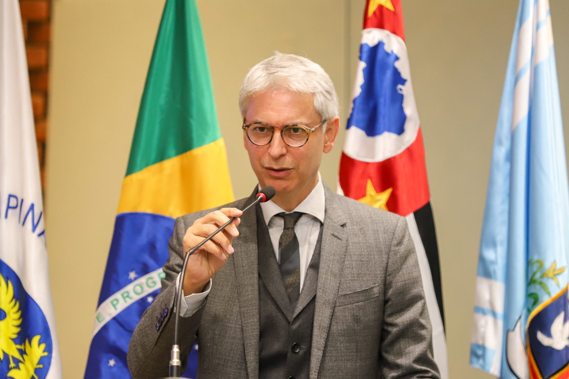 "procurador Ronaldo Lira está falando ao microfone no púlpito, com as bandeiras ao fundo"