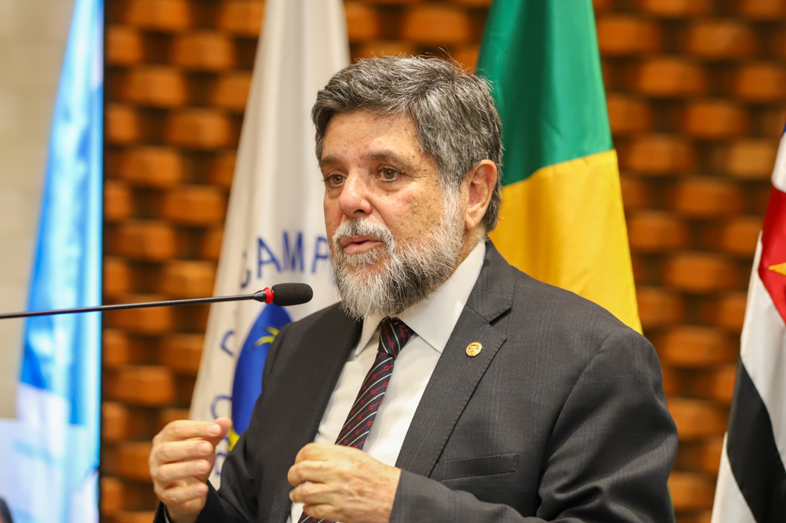 "Ministro Caputo Bastos fala ao microfone no púlpito, em frente a bandeiras"