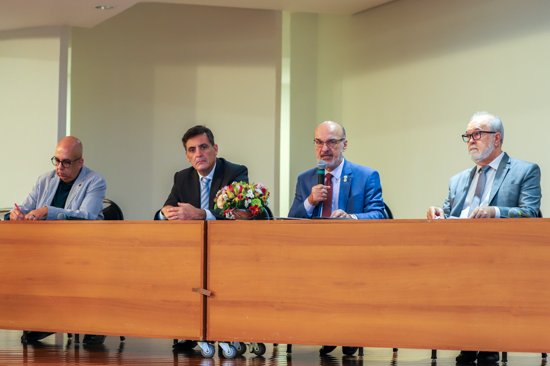 "Dr. Edmundo fala ao microfone na mesa alta com outros componentes do painel"