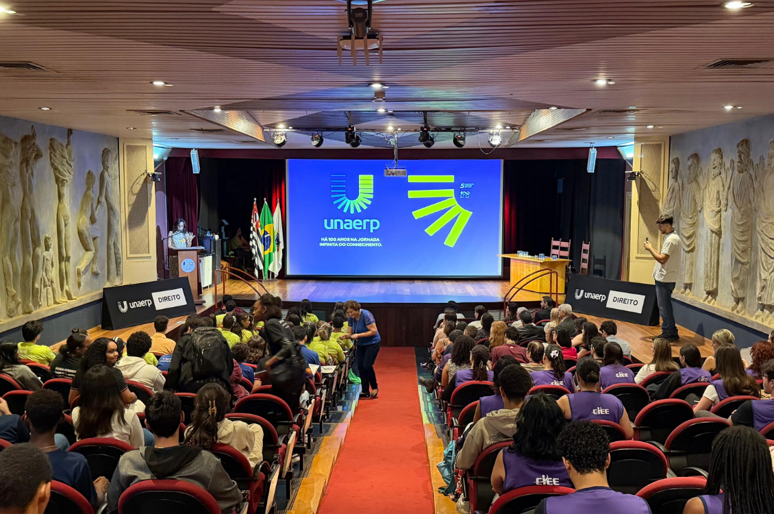 "doto geral do auditório da Unaerp lotado, com o telão azul ao fundo exibindo logotipo da universidade"