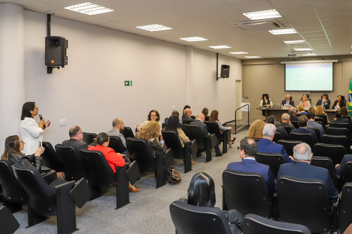 "foto geral do público com uma participante de pé falando ao microfone e os gestores sentados è mesa de trabalhos, ao fundo"