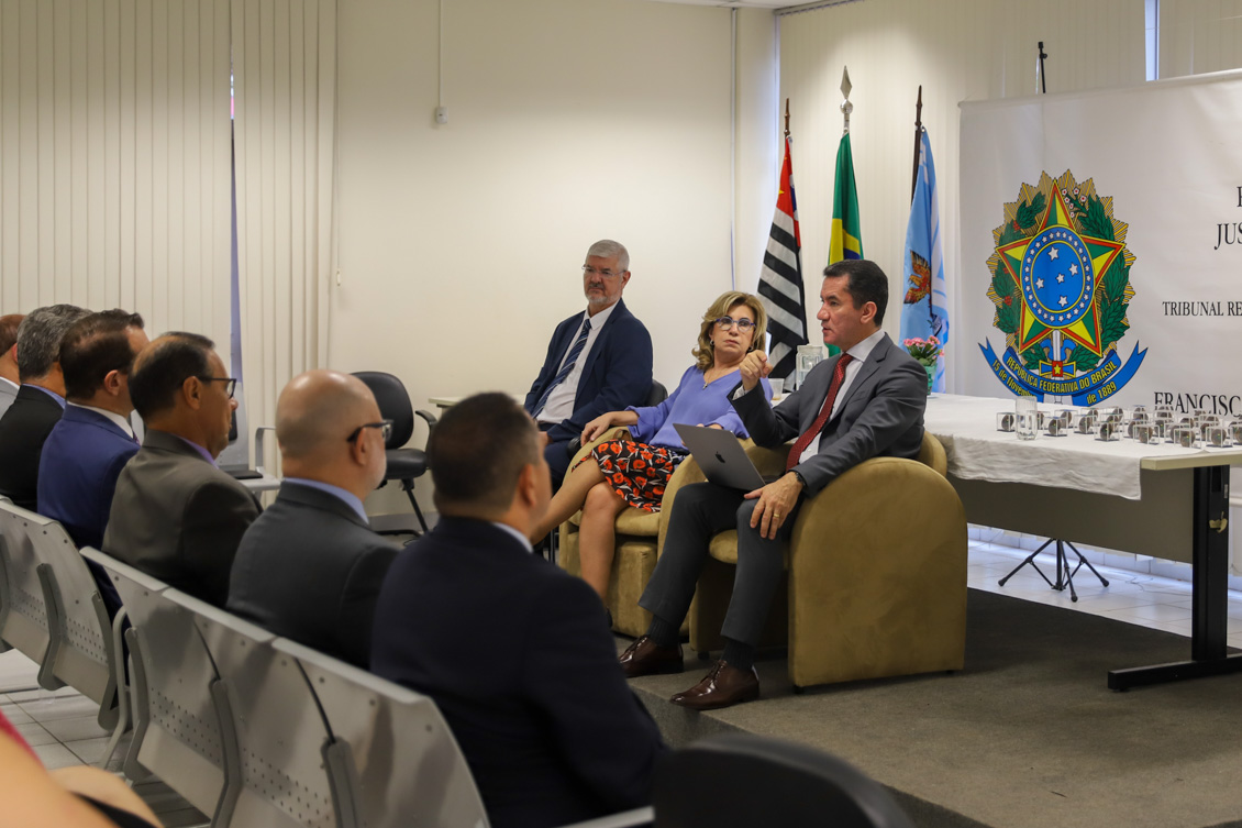 "Renan Fagundes fala ao microfone, ao lado de Ana Paula e Edison Pelegrini, sendo observado por parte dos juízes no auditório"
