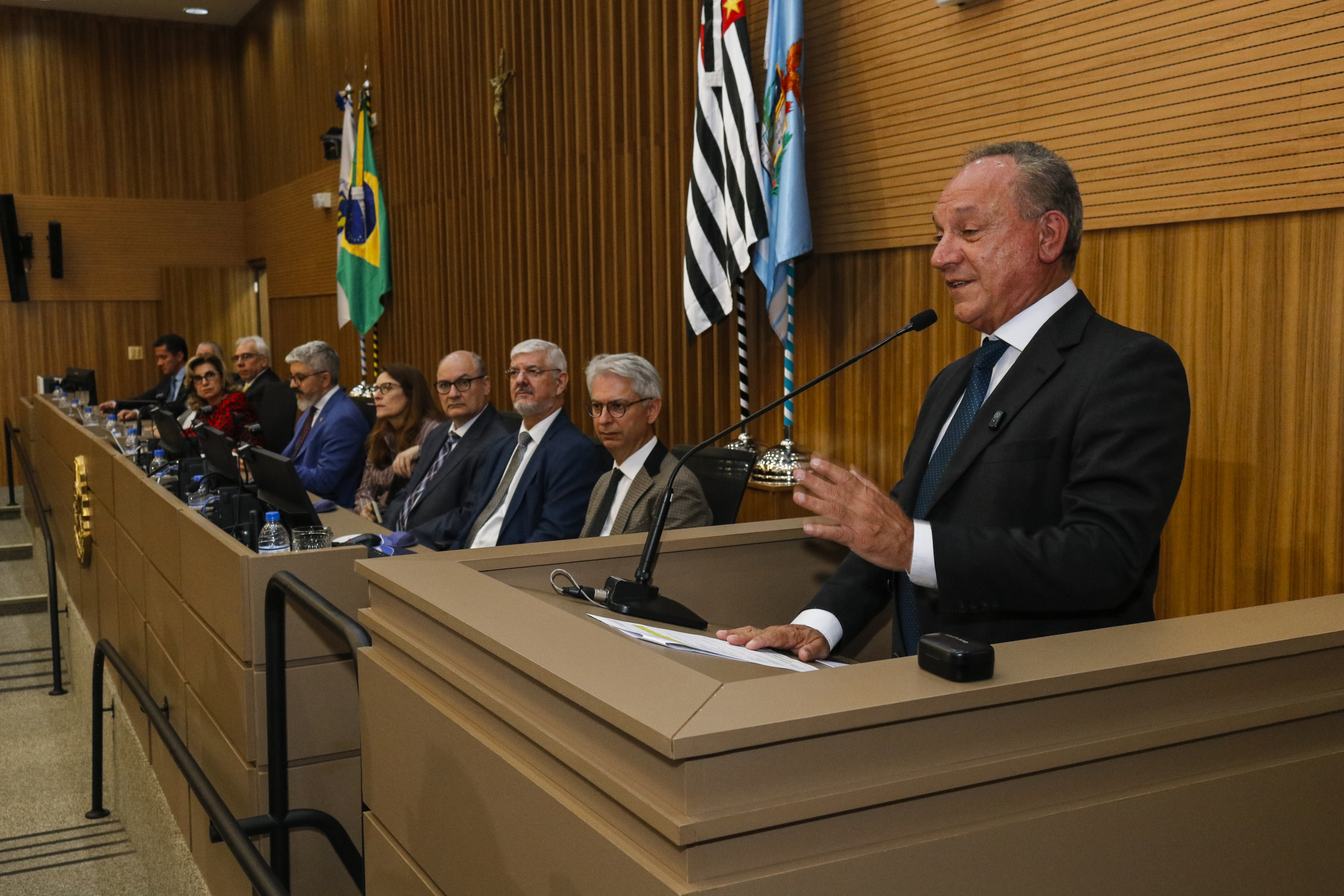"Vereador Rossini está no púlpito falando ao microfone, sendo observado pelas integrantes da mesa e público"
