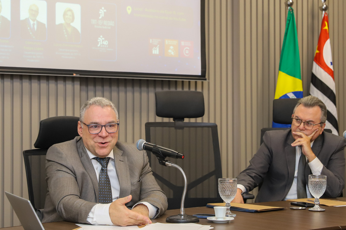 "Dois homens estão sentados. Um deles fala ao microfone"