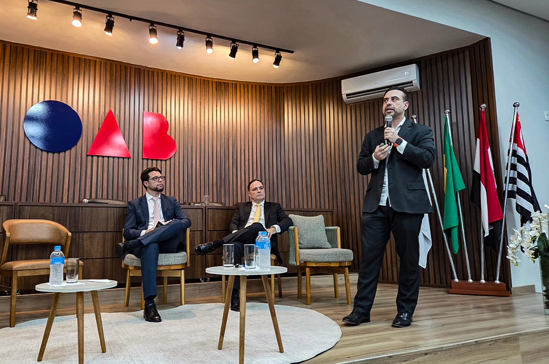 "Um homem está de pé, falando ao microfone, sendo observado por outros dois, que estão sentados em cadeiras de madeira no palco do auditório da OAB"