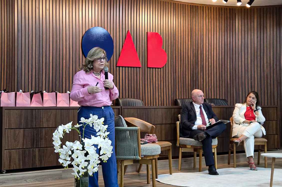 "Uma mulher fala ao microfone, em pé, sendo observado por um homem e uma mulher sentados em cadeiras de madeira no palco do auditório da OAB"