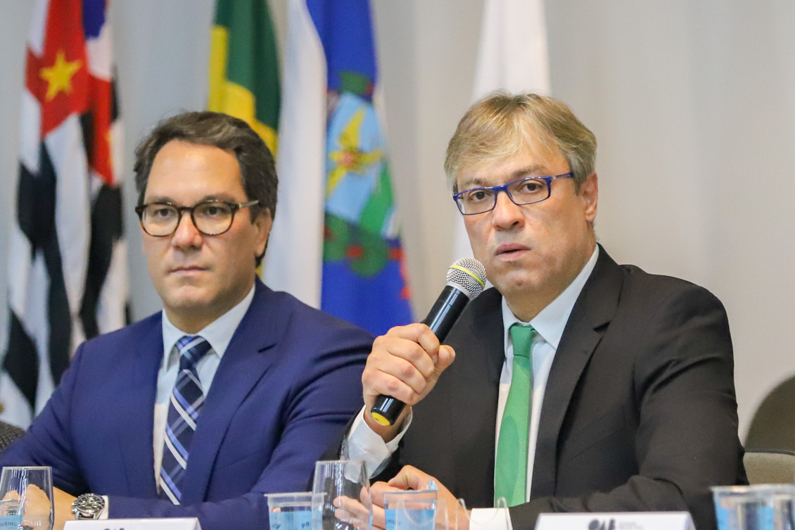 "imagem de dois homens sentados a mesa. Um está falando ao microfone"