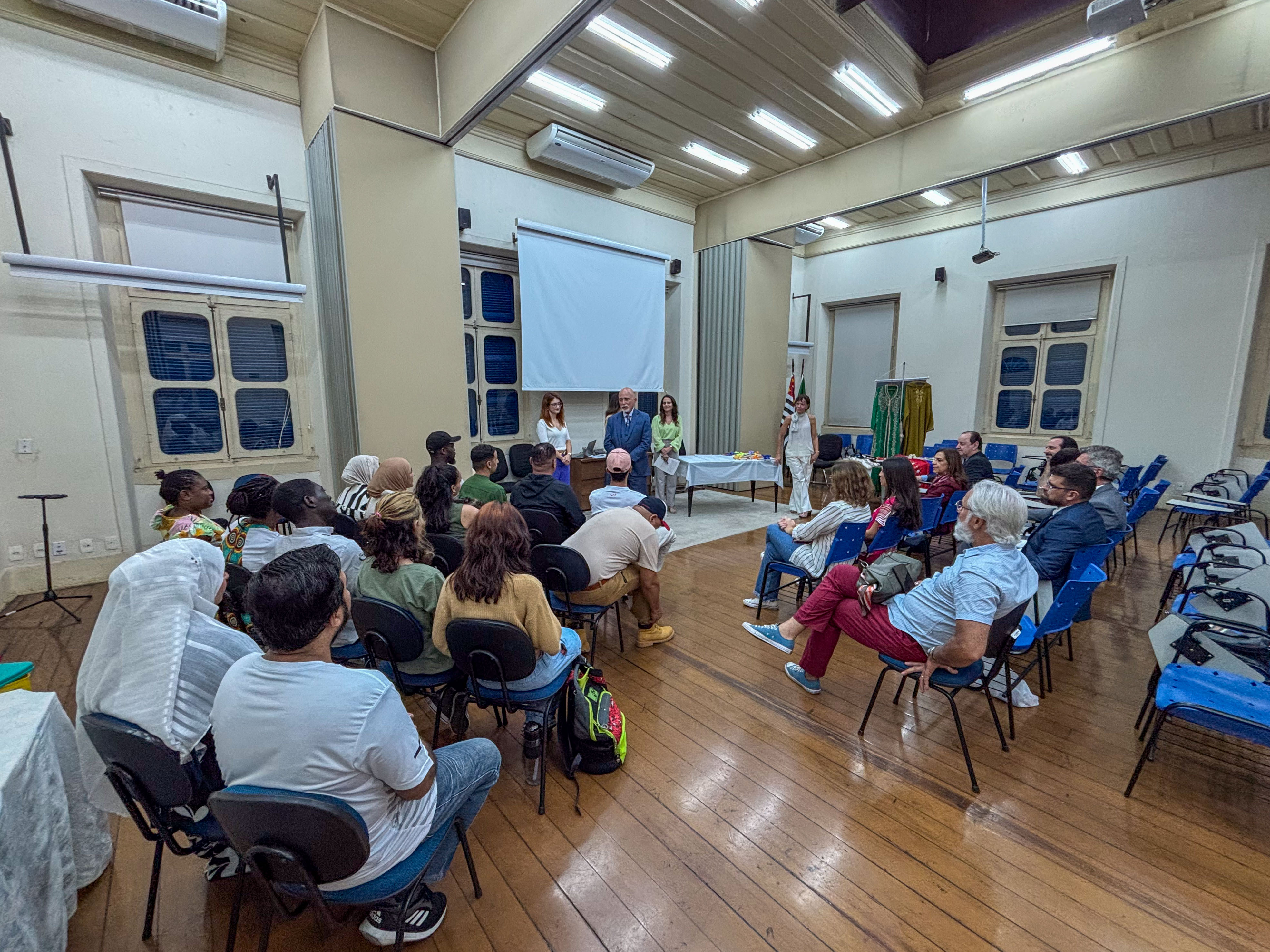 "imagem da turma de migrantes sentados escutando o desembargador Samuel Hugo Lima que fala à frente deles"