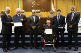 Servidor Sisenando ao centro, na cadeira de rodas, segurando certificado ao lado do juiz Guilherme Feliciano com o prêmio nas mãos. Ainda na foto, dirigentes do TST