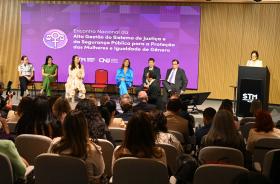 seis componente da mesa alta estão sentados em cadeiras dispostas no palco. Uma mulher fala ao microfone no púlpito, sendo observada pelo público.