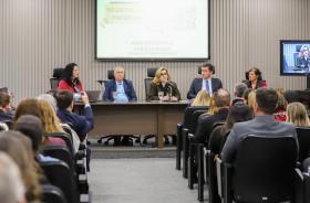 mesa de trabalhos composta pela Iara Gomes, Dr. Helcio, Dra. Ana Paula ao microfone, Dr. Renan e Dra. Regina. Todos sentados com o público em primeiro plano.