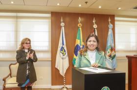 juíza está atrás de dispositivo, olhando para câmera após assinatura de termo de posse. À esquerda da tela, Dra. Ana Paula aplaude o ato.