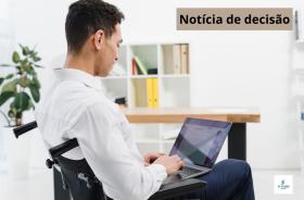 A foto retrata um rapaz sentado na cadeira de roda, digitando em um notbook.