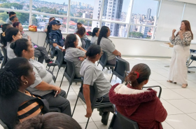 Mulheres estão sentadas olhando a palestrante que está em pé falando.