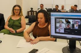 Mulher assina um papel que está sobre a mesa, sendo observada por outra, e com trabalhadores ao fundo, sentados. No lado direito da tela, um monitor com pessoas on-line