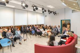 foto geral do auditório com foco no público, à direita da tela, os palestrantes estão sentados em um sofá vermelho