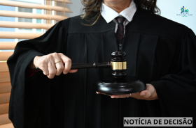 A imagem mostra uma pessoa vestindo uma toga preta, típica do Poder Judiciário. A pessoa segura um grande martelo de juiz: com a mão direita, apoia o martelo sobre uma base de madeira escura; com a mão esquerda, segura o cabo do martelo. A pessoa usa uma camisa branca com gravata escura sob a toga, e parte do seu cabelo castanho aparece sobre os ombros. Ao fundo, há persianas de madeira e um ambiente interno iluminado. No canto superior direito, aparece o selo dos “40 anos do TRT da 15ª Região – Campinas”. 