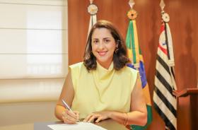 juíza Iara assina o termo de posse que está sobre um dispositivo de madeira. Ao fundo, três bandeiras.