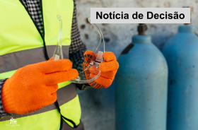 homem com colete reluzente e luvas laranjas segura óculos de proteção. Ao fundo, aparecem dois cilindros