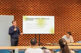 Thiago Rosa da Educorp está de pé com microfone na mão ao lado de uma tela que traz informações sobre linguagem simples. Em primeiro plano, os participantes do encontro acompanham a explanação