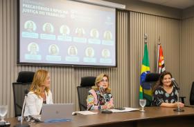 Mesa de encerramento com a Dra. Ana Paula no centro, tendo a Dra. Daniela de seu lado direito e a servidora Samira, do lado esquerdo. Ao fundo, o telão temático e bandeiras