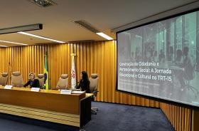 Dra. Ana Paula e Renata estão sentadas à mesa de trabalhos do Coleprecor, de frente de microfones, e ao lado, um telão exibe o tema da palestra