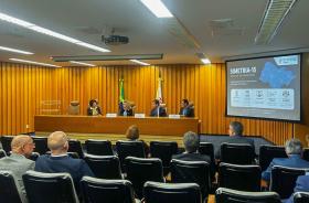 À frente da sala, quatro autoridades estão sentadas atrás de uma mesa de madeira, diante das bandeiras do Brasil e do Estado de São Paulo. À direita, um telão exibe slide do projeto apresentado pelo TRT-15. Na plateia, participantes acompanham a exposição sentados em cadeiras pretas dispostas em fileiras. O ambiente possui iluminação clara e paredes revestidas em madeira.