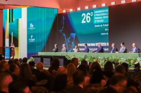A imagem mostra um grande auditório durante um congresso jurídico, visto a partir da plateia.  No palco, há uma longa mesa revestida de verde, decorada na parte frontal com arranjos de plantas. Oito pessoas estão sentadas atrás da mesa, aparentemente compondo uma mesa de autoridades ou palestrantes. À esquerda do palco, uma mulher está em pé diante de um púlpito, aparentemente falando ao público.  Ao fundo, grandes painéis de LED exibem uma identidade visual em tons de azul, turquesa e verde, com elementos 