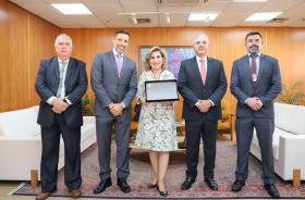 Cinco pessoas posam para a foto, em pé. Ao meio está uma mulher segurando uma placa.