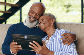 Foto de dois idosos abraçados sentados. Eles seguram um tablet e sorriem.