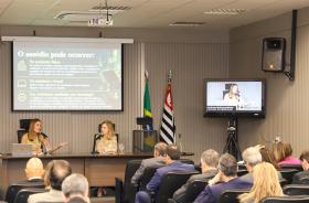 Dra. Daniela e Dra. Antonia na mesa de trabalhos com público em primeiro plano e telão ao fundo
