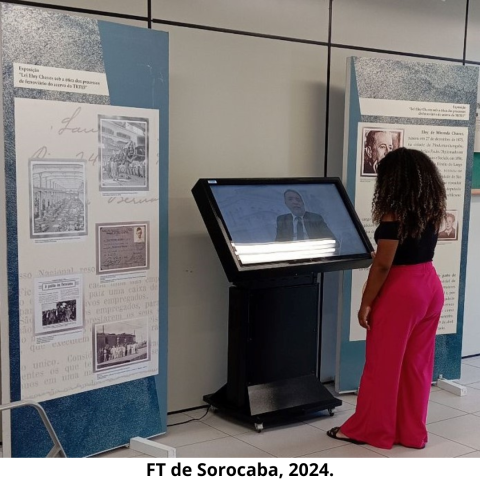 Mulher ao lado direito, de cabelos longos escuros, vestida com calça na cor vermelha e blusa preta, observa tela em que tem imagem de um homem, aos lados direito e esquerdo da tela, há dois painéis, com fundo verde e bege, textos e imagens. Atrás dos painéis, há uma parede na cor bege.