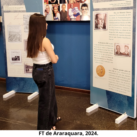 Mulher ao lado esquerdo, de cabelos longos escuros, vestida com calça na cor escura e blusa branca observa dois painéis, um ao lado direito e outro ao esquerdo, ambos com textos e imagens. Atrás dos painéis, há uma parede na cor azul.