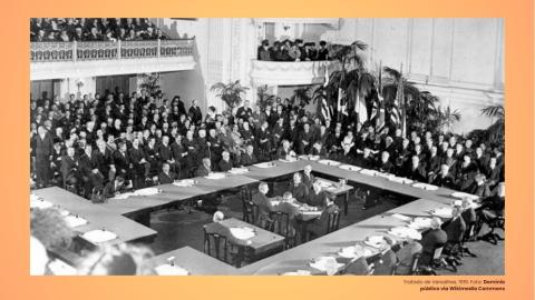 Fotografia mostra homens, brancos, vestidos de ternos e reunidos ao redor de uma mesa quadrada. Quase não há mulheres na sala. Trata-se do Tratado de Versalhes, em 1919. 