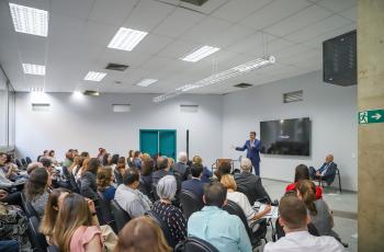 Ao fundo, Luiz Fernando fala ao microfone, sendo observado por doutor Samuel, sentado e demais participantes.