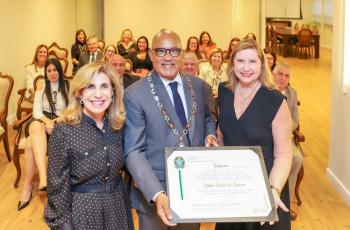 Dr. Fábio com o diploma e o colar, ladeado pela presidente Dra. Ana Paula e Dra. Rita. Ao fundo, o público sentado.