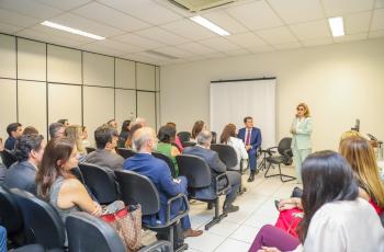 Dra. Ana Paula está no canto direito da tela, gesticulando, sendo observada pelos juízes sentados no auditório. 