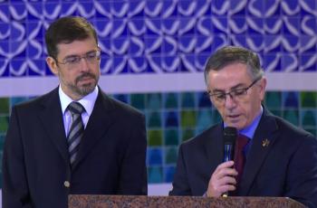 Dr. João Batista está no púlpito, com microfone na mão, fazendo a leitura da carta,  e sendo observado pelo Dr. Adhemar.