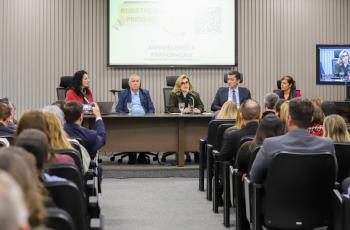 mesa de trabalhos composta pela Iara Gomes, Dr. Helcio, Dra. Ana Paula ao microfone, Dr. Renan e Dra. Regina. Todos sentados com o público em primeiro plano.