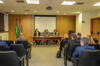 Mesa de trabalhos composta pela Dr. Marcello, Dra. Ana Paula, que está ao microfone, e Dra. Keity. Em primeiro plano, pessoas estão acompanhando a explanação, sentadas nas cadeiras do auditório
