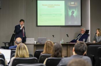 imagem geral do auditório com os palestrantes ao fundo e o público presente em primeiro plano. No telão, o tema da palestra