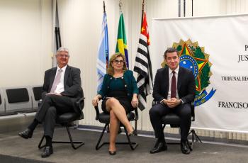 foto do Dr. Edison, Dra. Ana Paula e Dr. Renan, sentados em cadeiras sobre um tablado, com bandeiras ao fundo e um banner alusivo ao Fórum Trabalhista local