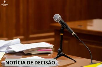 Fotografia colorida mostra o interior de uma sala de audiência. Em primeiro plano, há uma mesa de madeira com vários papéis, documentos e pastas empilhados. Sobre a mesa, um microfone prateado está fixado em um pedestal preto, direcionado para o local onde normalmente fala uma das partes ou testemunhas. No canto superior esquerdo da imagem, aparece o logotipo do TRT da 15ª Região – Campinas. Na parte inferior, uma faixa cinza contém o texto “NOTÍCIA DE DECISÃO” em letras maiúsculas pretas.