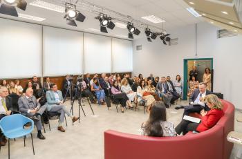 foto geral do auditório com foco no público, à direita da tela, os palestrantes estão sentados em um sofá vermelho