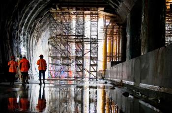 obra em túnel com três funcionários de pé e de costas, à esquerda, e imagens refletindo no chão molhado