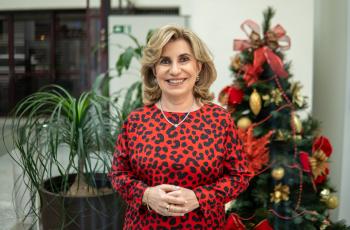 Dra. Ana Paula está de vermelho, num plano americano com fundo desfocado, em frente a uma árvore de natal, com mãos entrelaçadas