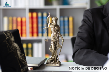 estante de livros ao fundo. No centro da mesa, a estátua da justiça. No lado esquerdo da tela, um notebook. No lado direito, imagem parcial de um homem de terno